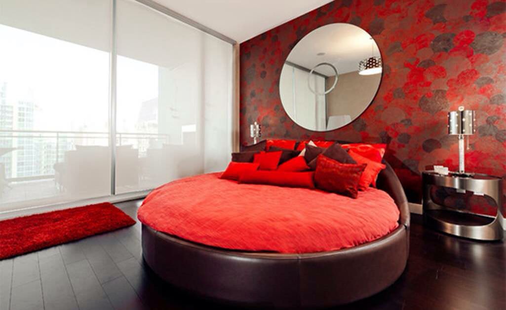 تخت خواب گرد قهوه ای سوخته با روتختی قرمز که در کنار آن آینه دایره ای و پاتختی بیضی برای ایجاد هارمونی استفاده شده است